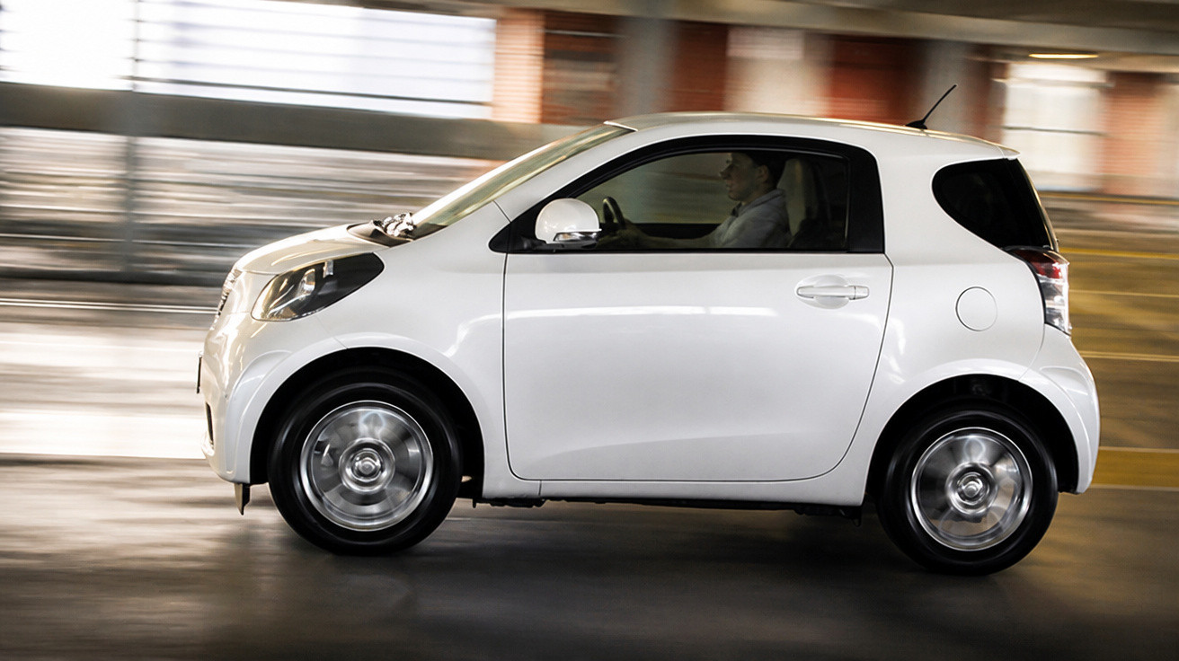 Carro branco compacto em movimento, com motorista, em ambiente urbano com fundo desfocado.