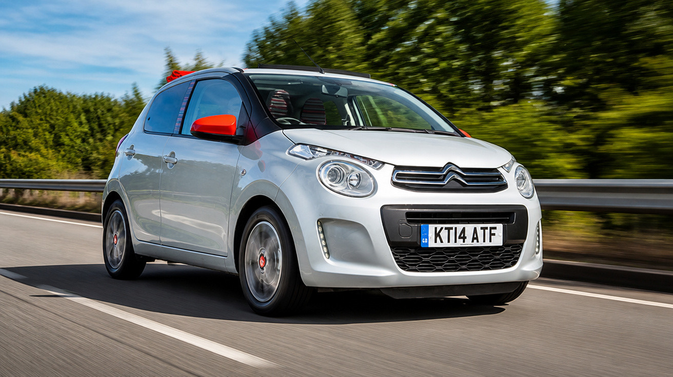 Carro Citroën branco com detalhes vermelhos em movimento em estrada asfaltada cercada por vegetação.