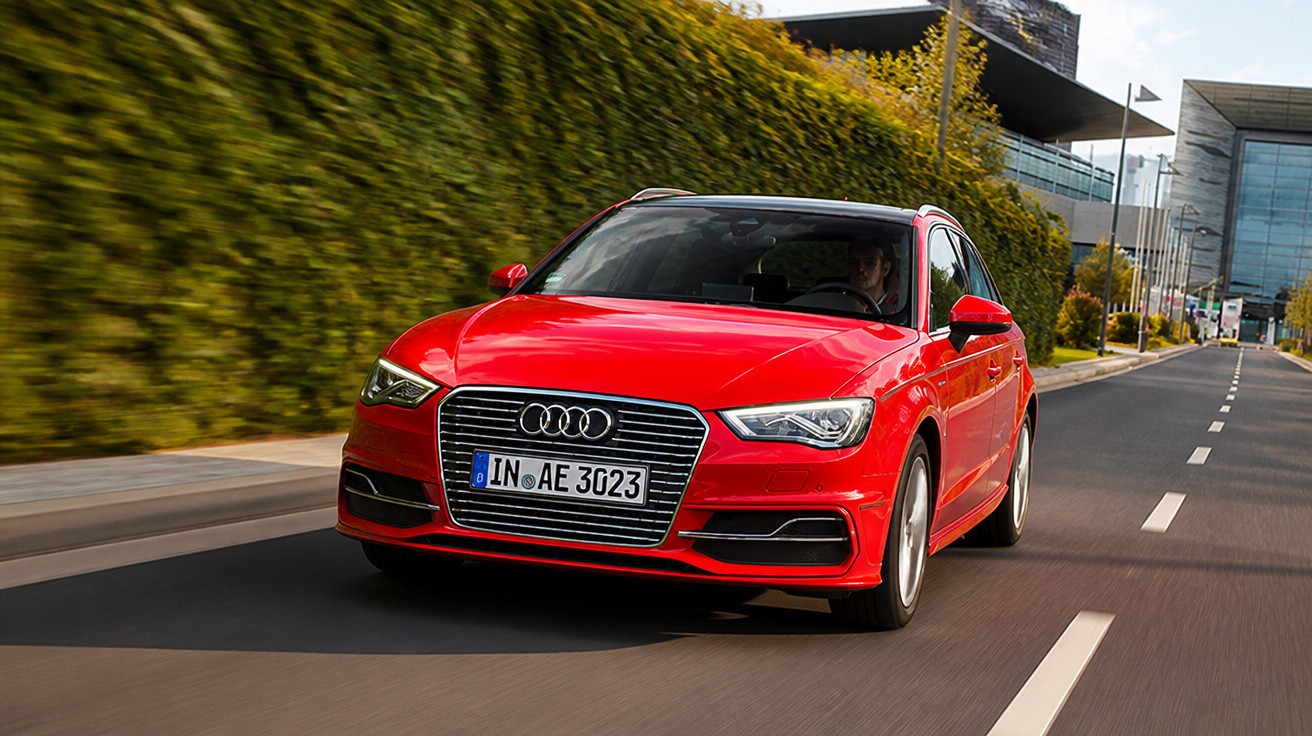 Carro Audi vermelho em movimento em estrada urbana com edifícios e vegetação ao fundo.