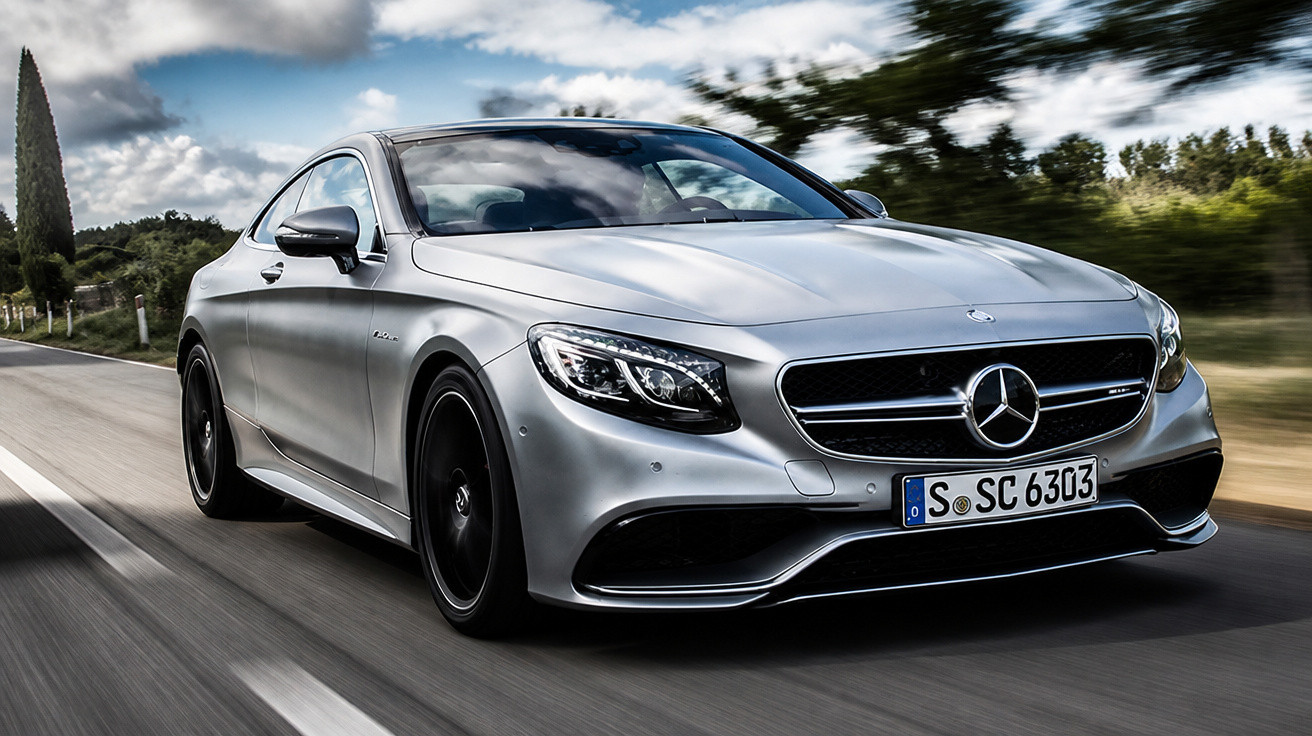 Carro esportivo Mercedes-Benz prata em movimento em estrada cercada por vegetação e céu nublado.