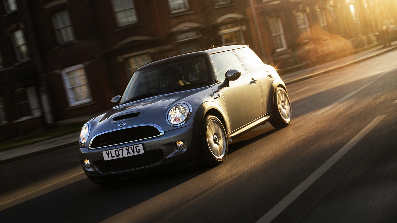 Carro Mini Cooper cinza escuro em movimento em uma rua urbana ao entardecer.