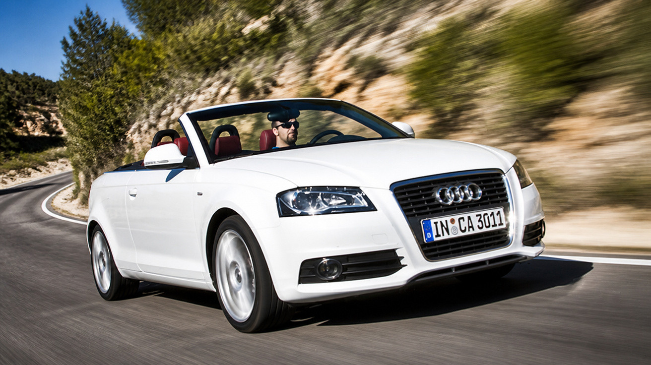 Carro conversível branco Audi com motorista usando óculos escuros em estrada sinuosa com paisagem natural.