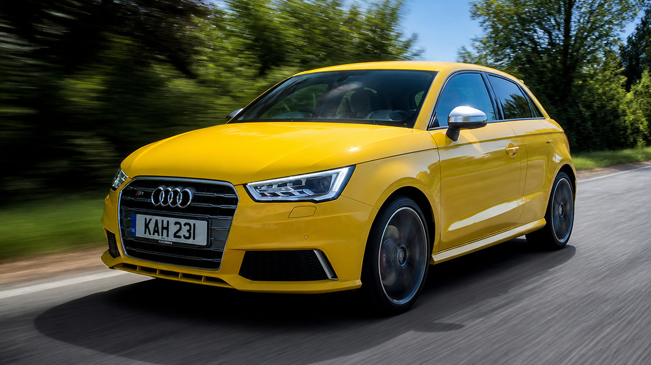 Carro hatchback amarelo Audi em movimento em estrada com árvores ao fundo.