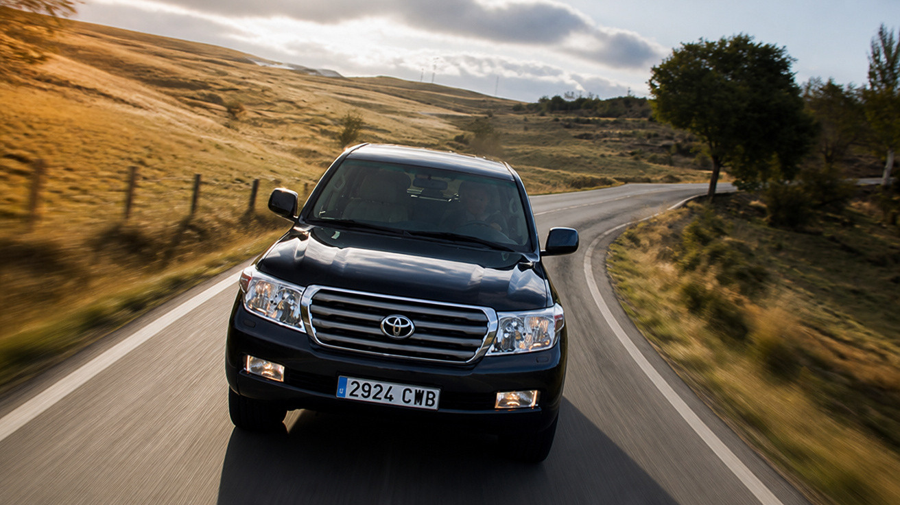 Carro preto Toyota Land Cruiser em estrada sinuosa cercada por campos e árvores ao entardecer.