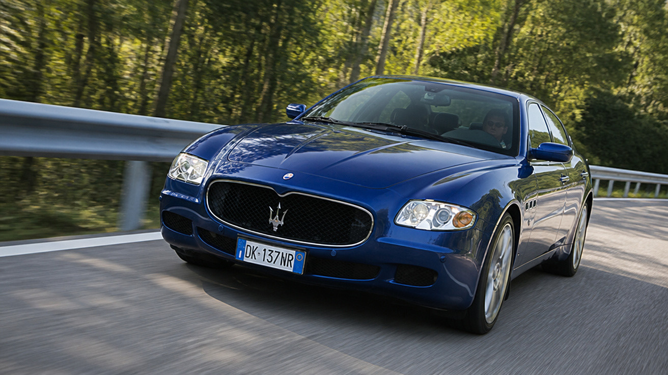 Carro esportivo azul Maserati dirigindo em estrada cercada por árvores e guardrail metálico.