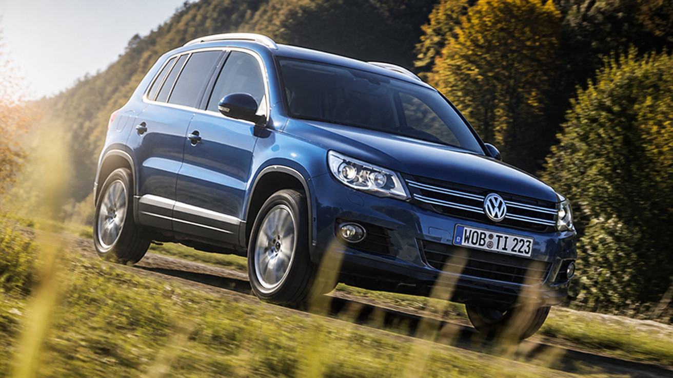 Carro SUV Volkswagen azul em estrada rural com paisagem de árvores e montanhas ao fundo.