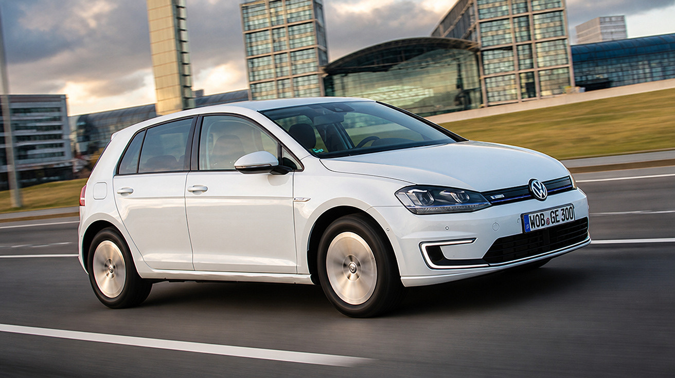 Carro branco Volkswagen Golf elétrico em movimento, com prédios modernos ao fundo em uma via urbana.