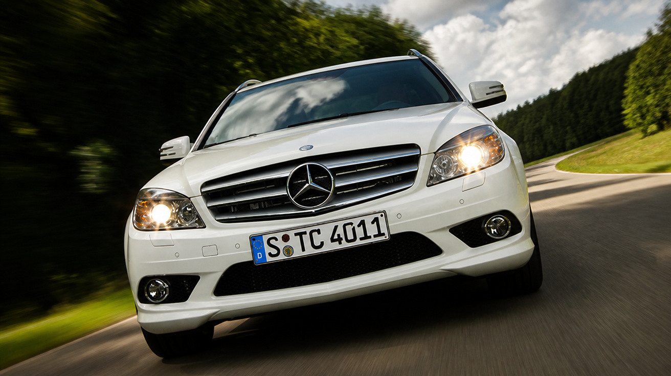 Carro Mercedes-Benz branco em movimento em estrada sinuosa com faróis acesos em dia nublado.