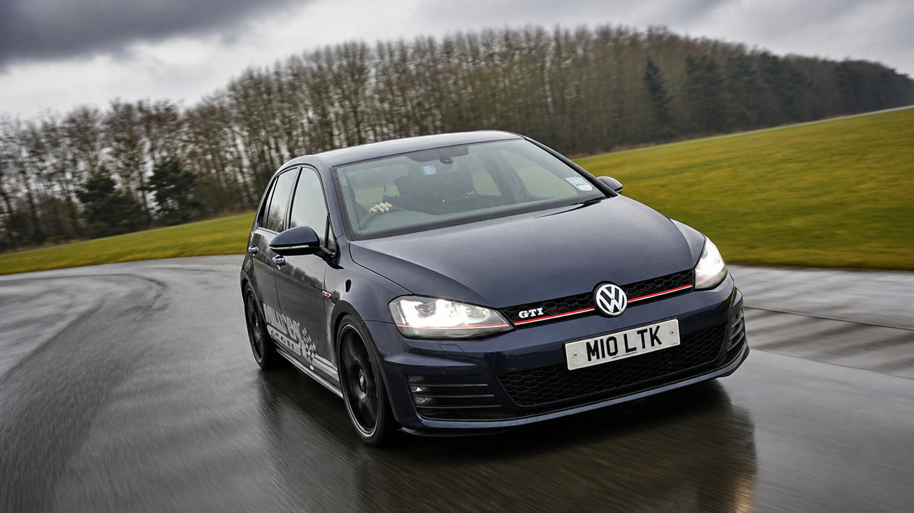 Carro Volkswagen Golf GTI preto com faróis ligados, em curva de pista molhada e fundo de árvores.