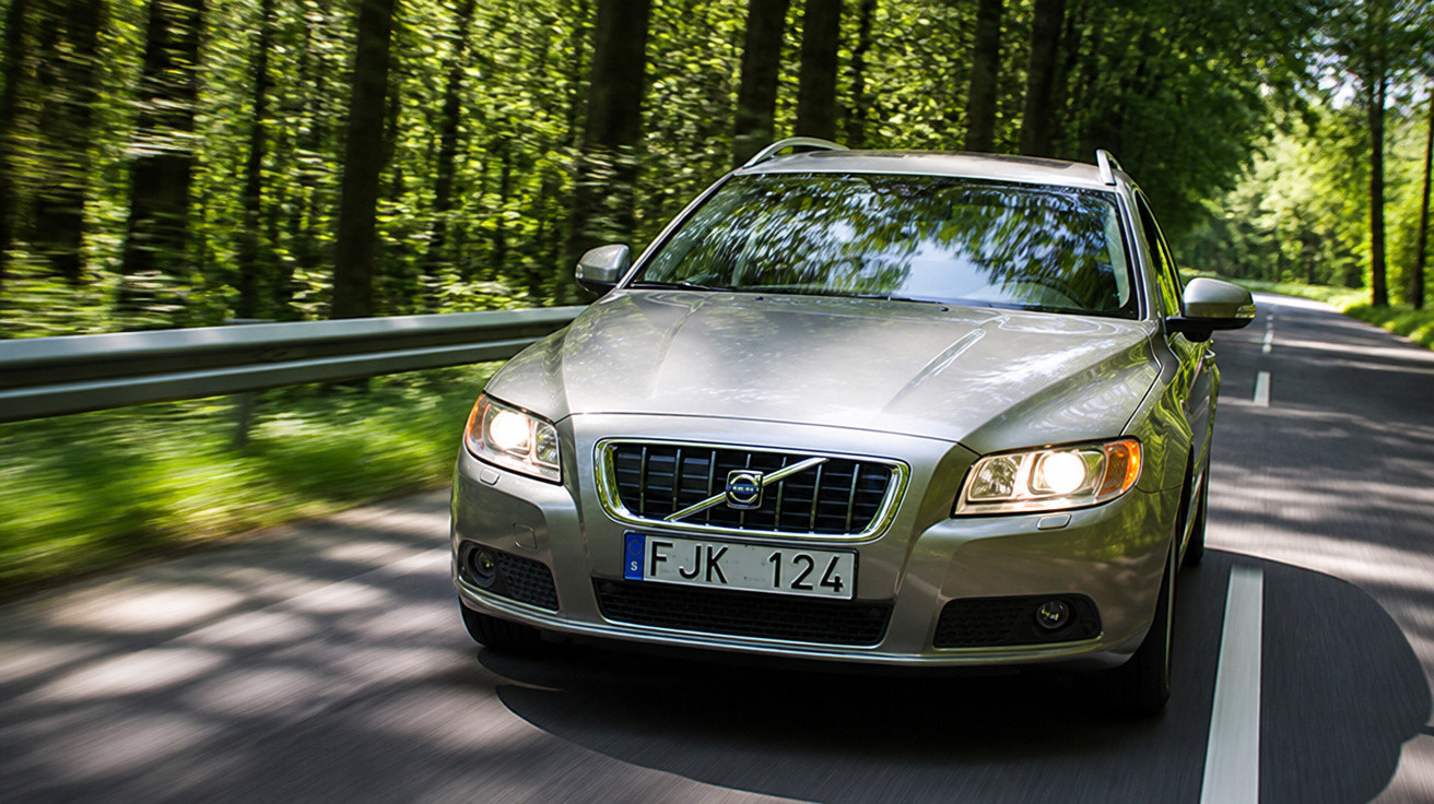 Carro prata Volvo em movimento em estrada cercada por árvores verdes em dia ensolarado.