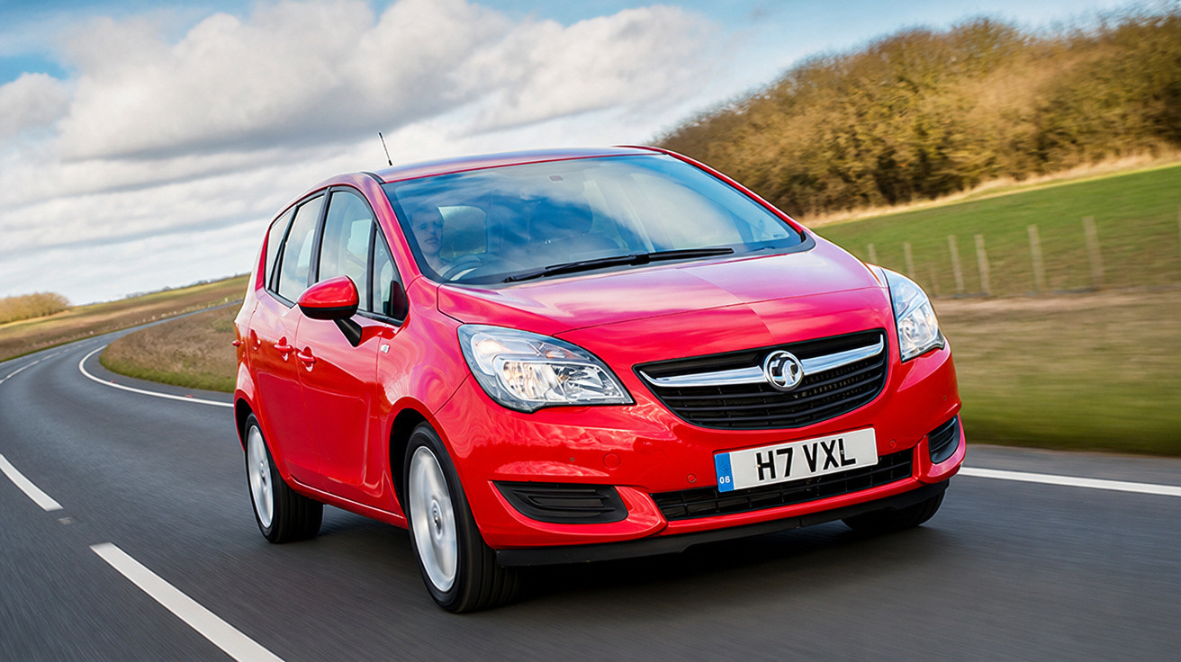 Carro hatchback vermelho trafegando em estrada curva com vegetação ao fundo em dia nublado.