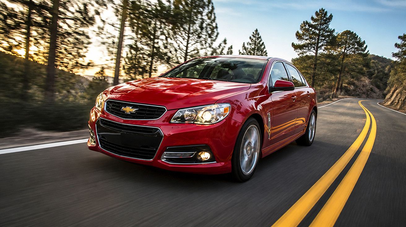 Carro vermelho Chevrolet circulando em estrada sinuosa cercada por árvores ao pôr do sol.