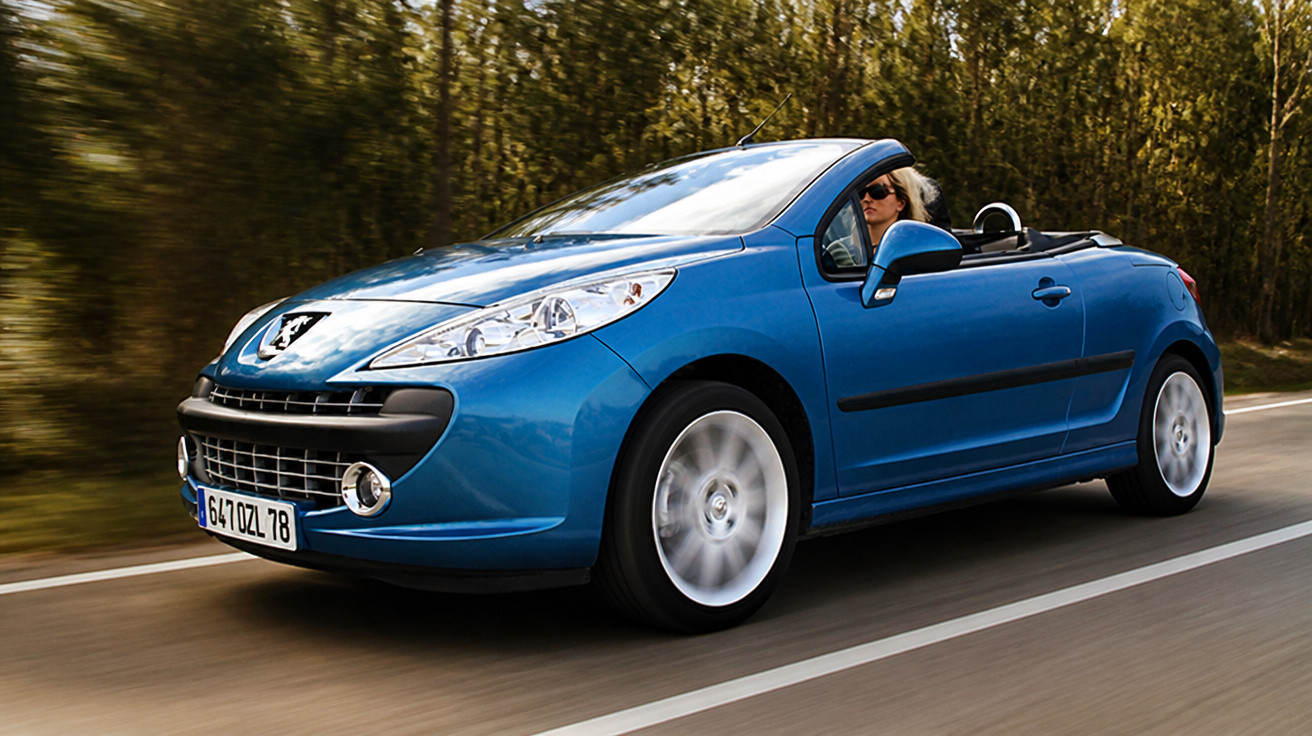 Carro conversível azul Peugeot em movimento em estrada cercada por árvores.