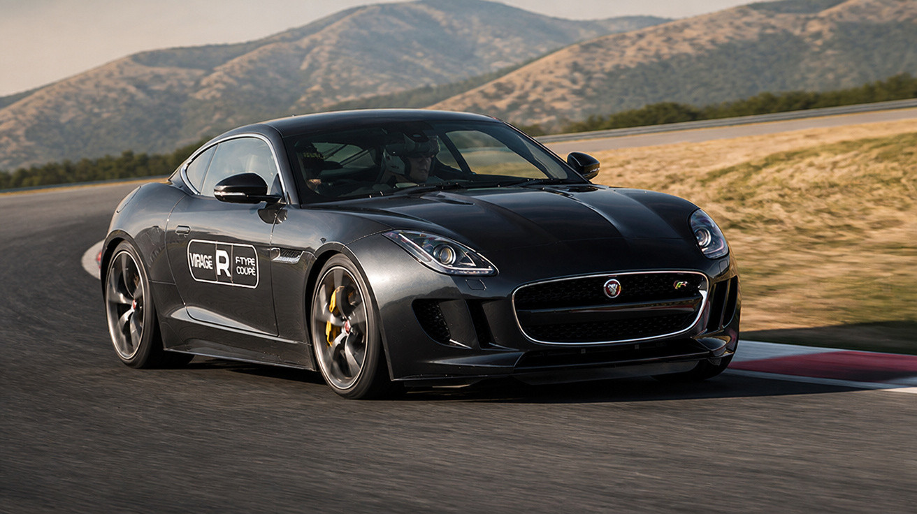Carro esportivo preto Jaguar F-Type Coupé em curva fechada na pista de corrida com montanhas ao fundo.