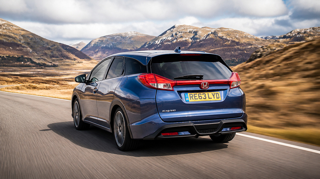 Carro hatchback azul Honda Civic circulando em estrada com montanhas ao fundo em dia nublado.