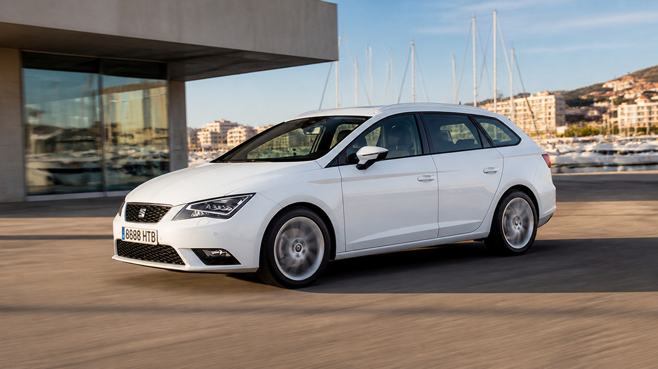 Carro branco modelo perua em movimento próximo a marina com veleiros ao fundo em dia ensolarado.