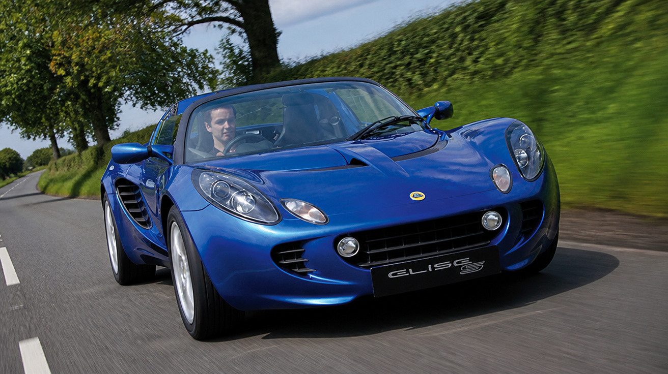 Carro esportivo azul Lotus Elise S em movimento em estrada cercada por árvores e vegetação.