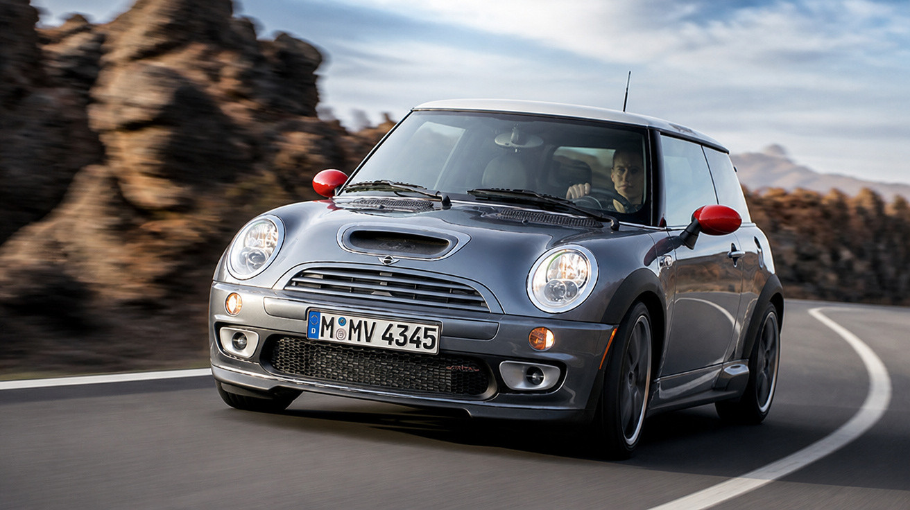 Carro Mini Cooper cinza com detalhes vermelhos trafegando em estrada sinuosa com pedras ao fundo.