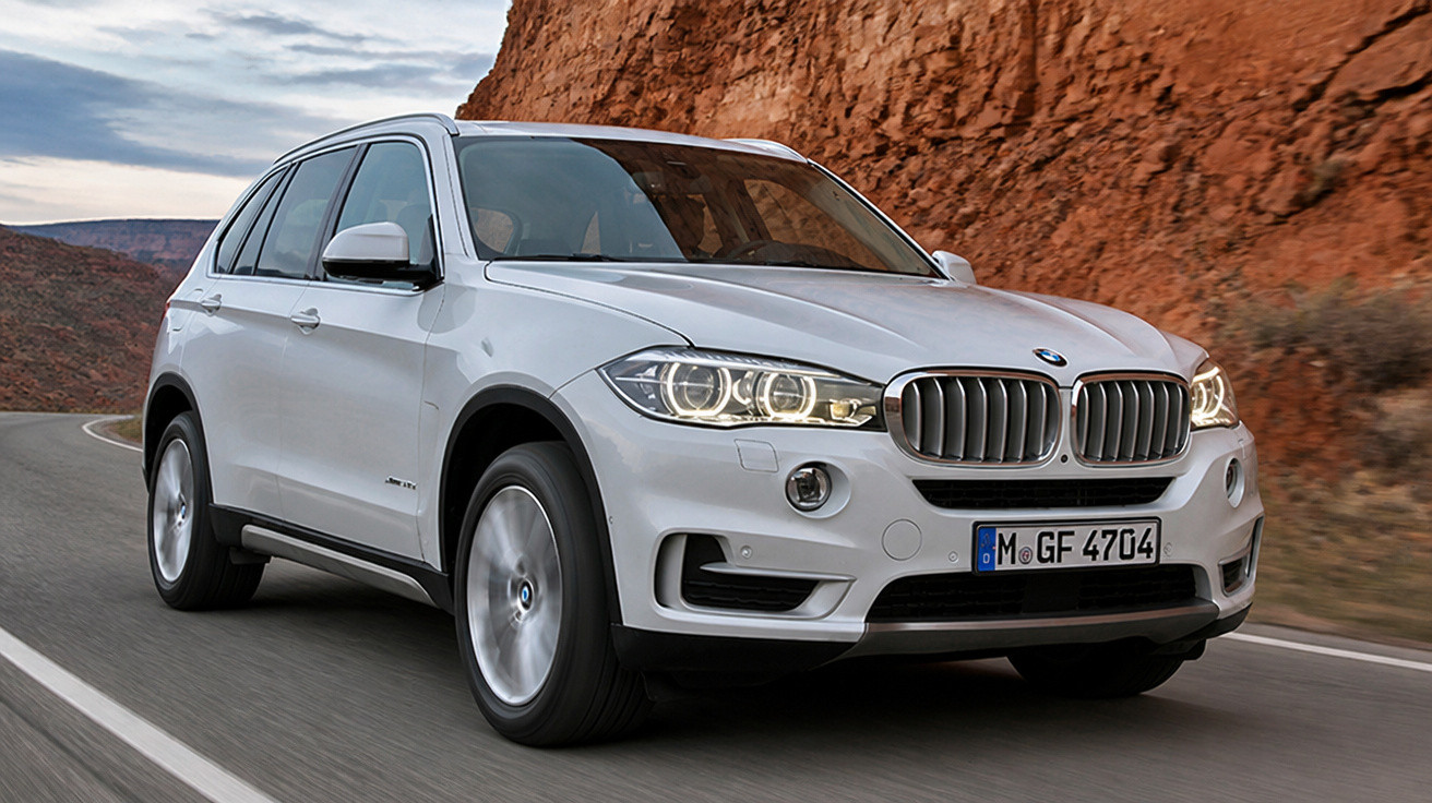 SUV branco BMW X5 em estrada sinuosa com paisagem rochosa ao fundo, luzes dianteiras acesas.