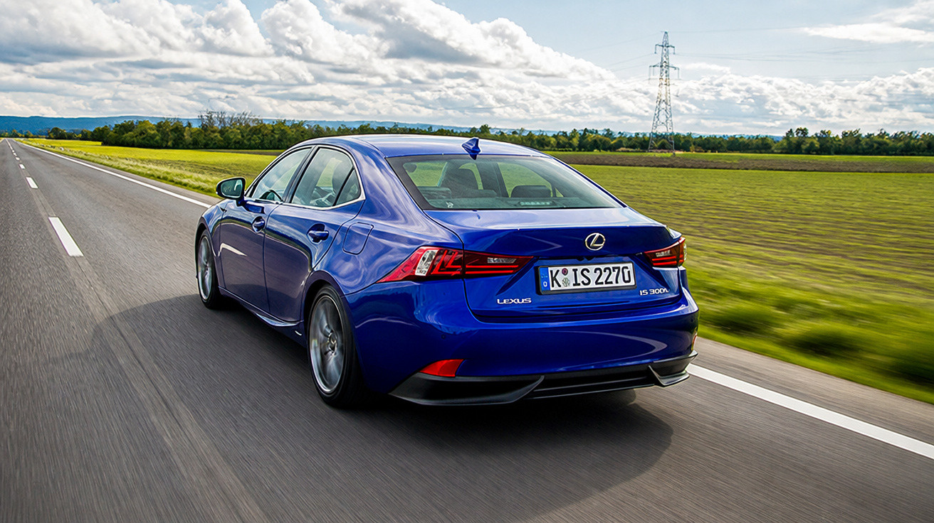 Carro sedã azul Lexus em estrada reta com céu nublado e campo verde ao redor.