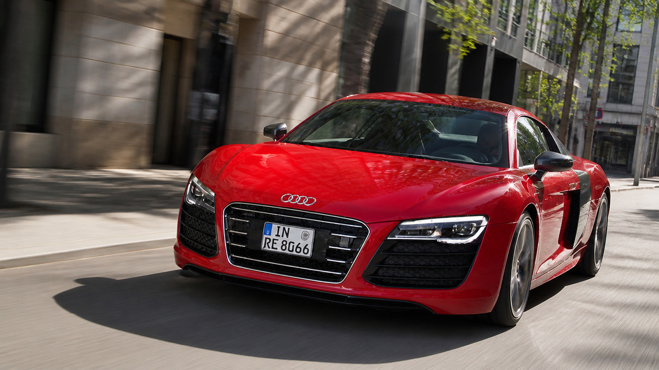 Carro esportivo Audi R8 vermelho circulando em rua urbana com prédios ao fundo.