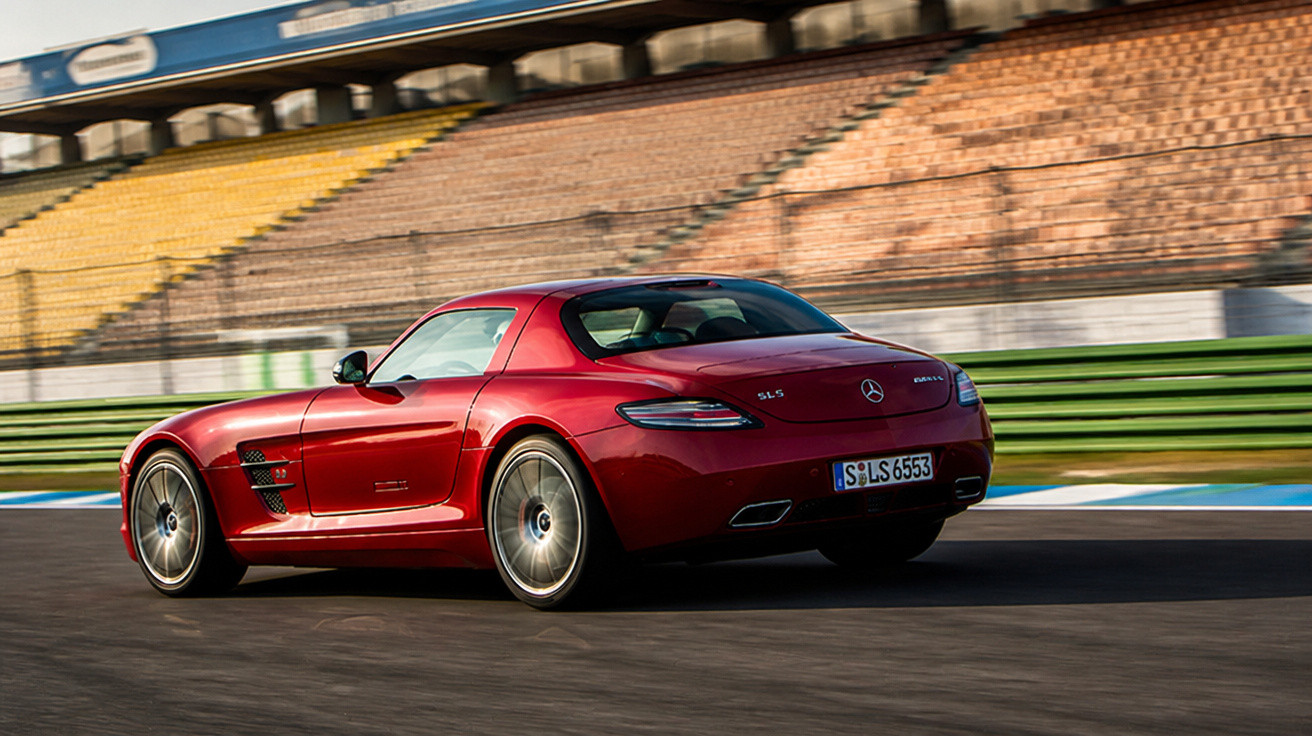 Mercedes-Benz SLS AMG vermelho em alta velocidade em pista de corrida com arquibancada ao fundo.