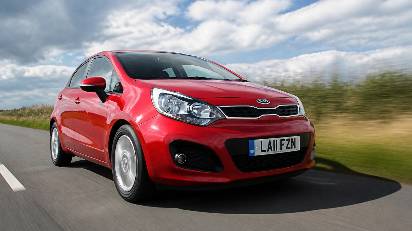 Carro vermelho Kia hatchback em movimento numa estrada com céu nublado ao fundo.