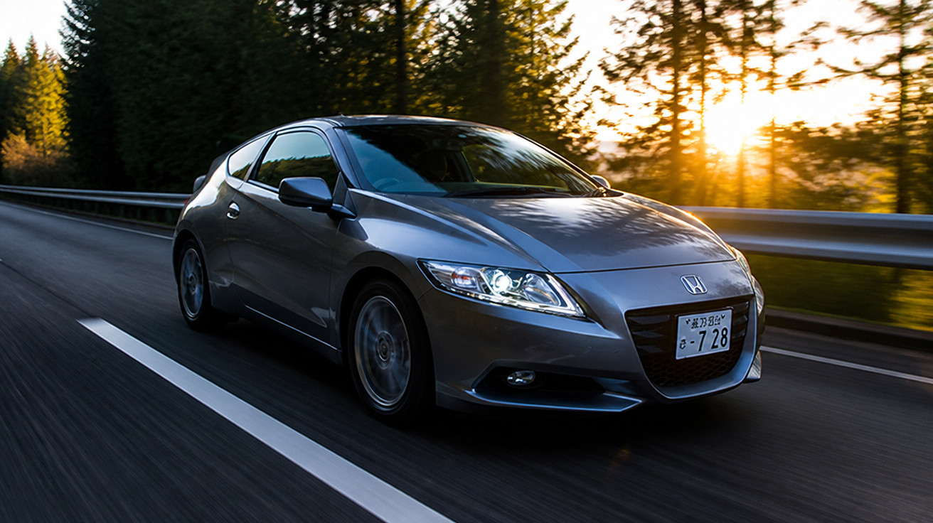 Carro cinza em movimento na estrada com árvores e pôr do sol ao fundo.