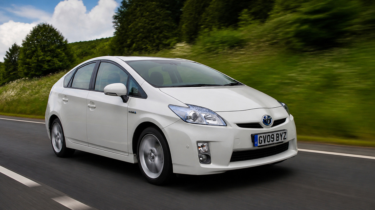 Carro branco Toyota Prius híbrido em estrada cercada por árvores e céu parcialmente nublado.