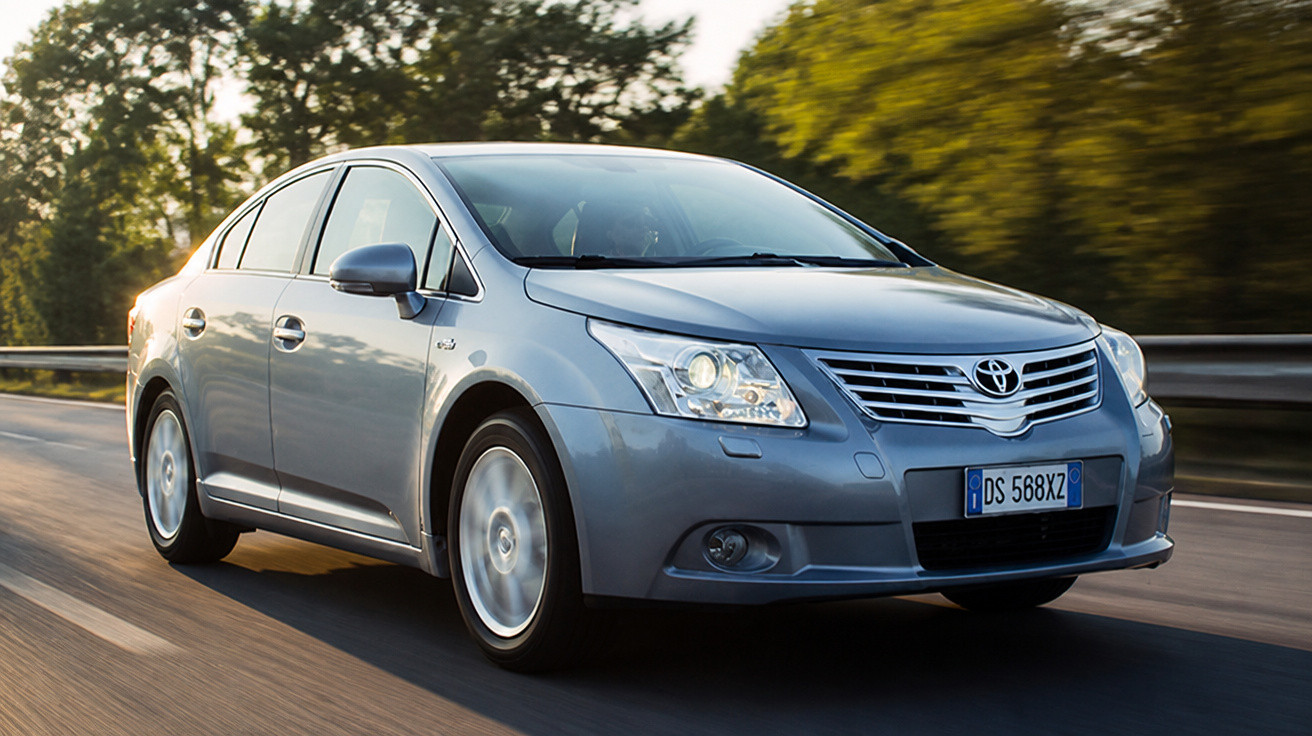 Carro sedan cinza Toyota em movimento em estrada cercada por árvores ao anoitecer.