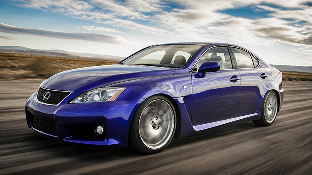Carro sedã azul da marca Lexus em movimento em estrada deserta com céu nublado ao fundo.