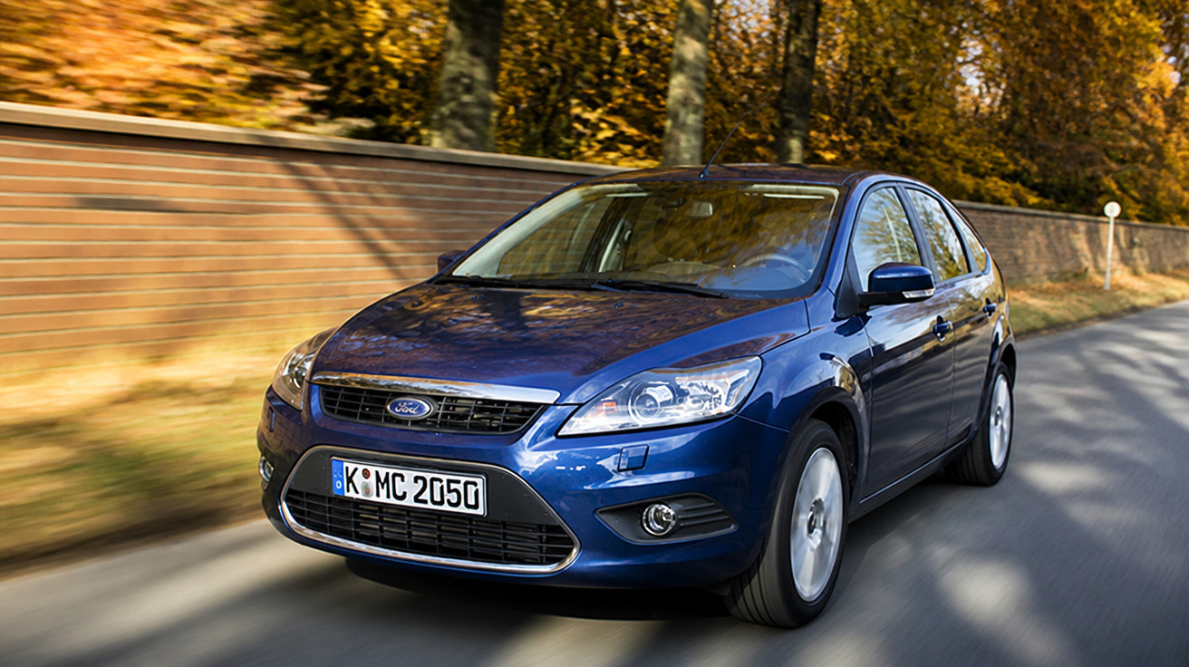 Carro azul Ford Focus em movimento em estrada com árvores e muro ao fundo.