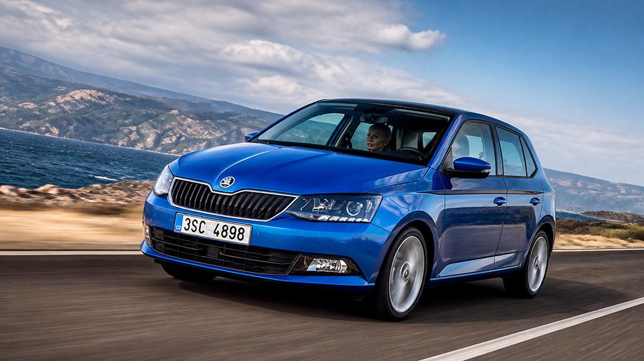 Carro hatchback azul modelo Skoda circulando em estrada costeira com montanhas ao fundo.