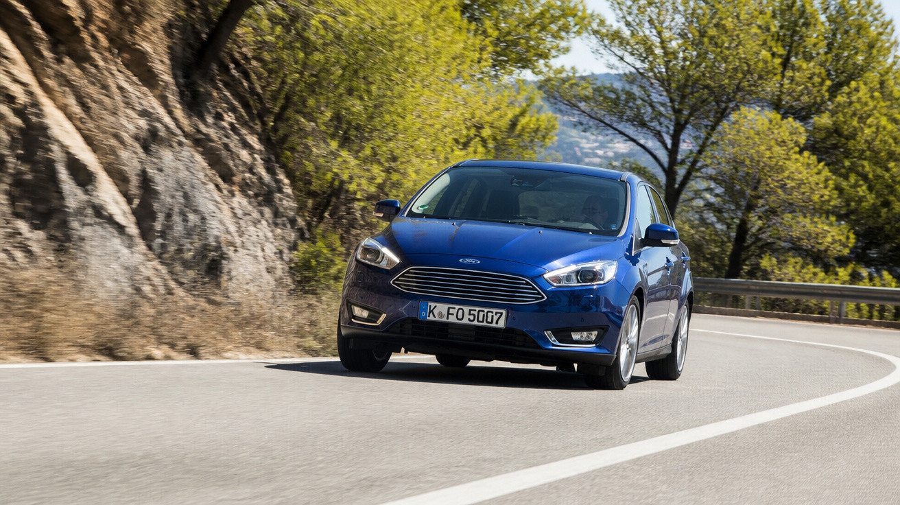 Carro azul Ford Focus na curva de estrada cercada por árvores e rochas em dia ensolarado.