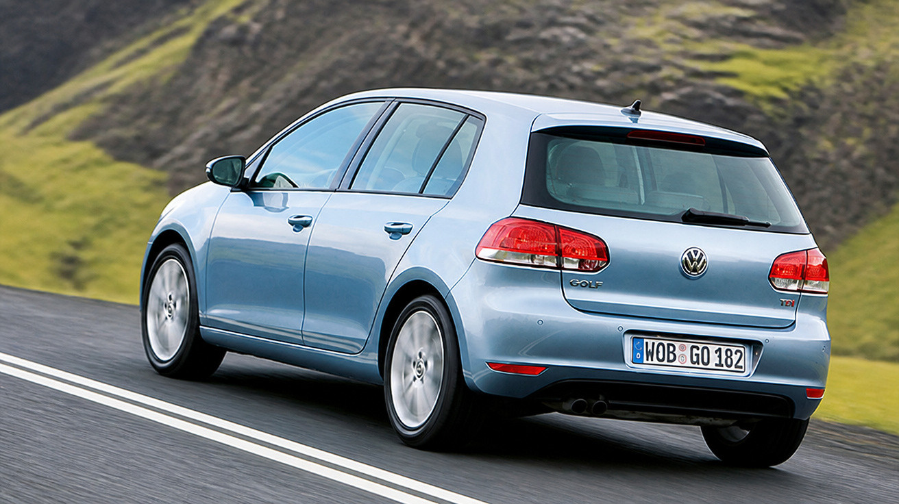 Carro Volkswagen Golf azul em estrada asfaltada com colinas verdes ao fundo em dia claro.
