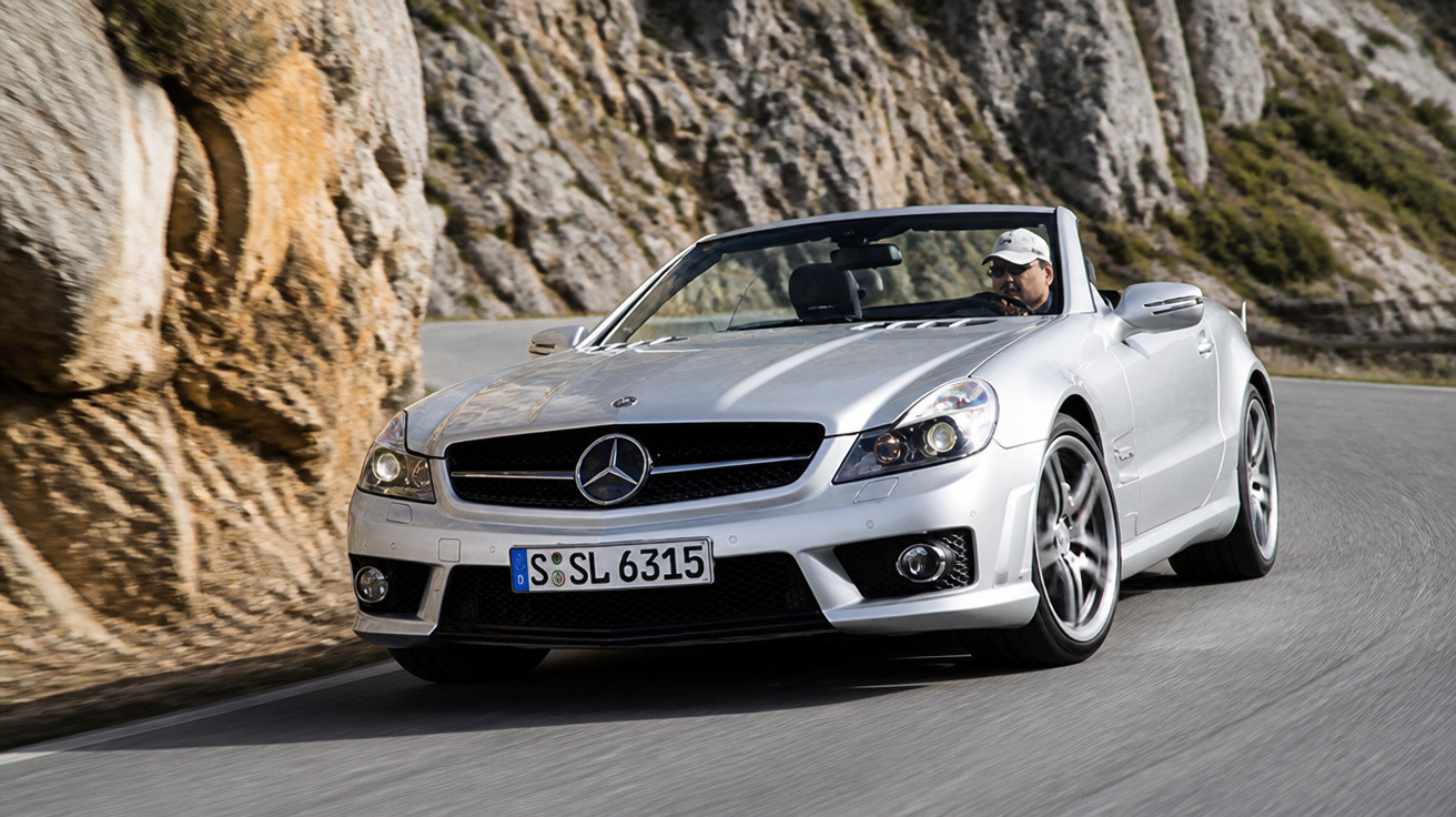 Carro conversível Mercedes-Benz prata em curvas de estrada montanhosa com motorista usando boné branco.