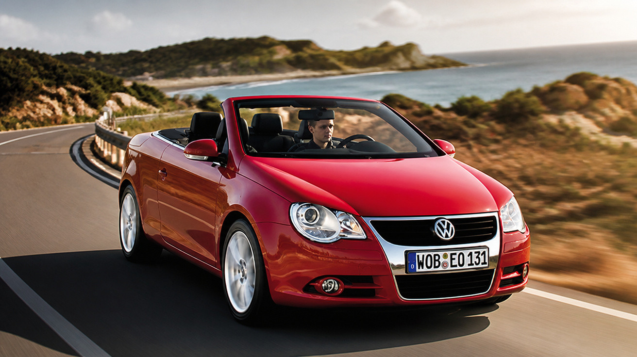 Carro conversível vermelho Volkswagen em estrada costeira com motorista, paisagem com colinas e mar ao fundo.